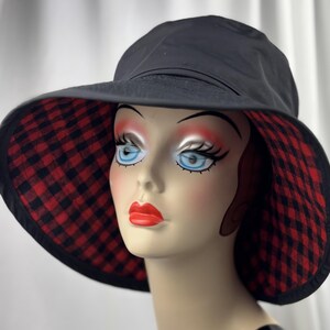 Pu&ograve; includere: Cappello da sole nero con tesa larga. L'interno della tesa presenta un motivo a quadri rossi e neri. Il cappello &egrave; esposto su una testa di manichino con occhi blu e rossetto rosso.