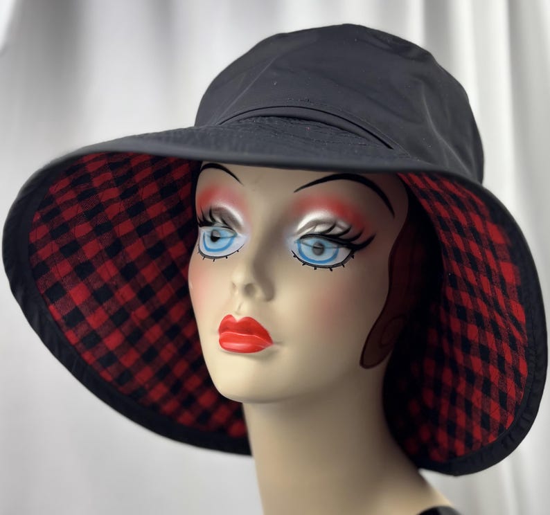 Pu&ograve; includere: Cappello da sole nero con tesa larga. L'interno della tesa presenta un motivo a quadri rossi e neri. Il cappello &egrave; esposto su una testa di manichino con occhi blu e rossetto rosso.