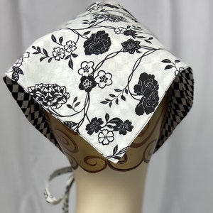 May include: A white and black floral patterned bonnet. The bonnet features a repeating pattern of black flowers and vines on a white background. The edges are trimmed with a black and white checkered pattern. The bonnet is tied with a matching fabric.