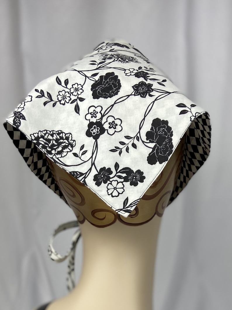 May include: A white and black floral patterned bonnet. The bonnet features a repeating pattern of black flowers and vines on a white background. The edges are trimmed with a black and white checkered pattern. The bonnet is tied with a matching fabric.