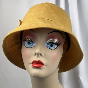 Puede incluir: Un sombrero de pescador color amarillo mostaza con un ala ancha inclinada hacia abajo y un peque&ntilde;o detalle de lazo decorativo en el lateral. El sombrero est&aacute; confeccionado en una tela suave similar a la gamuza y se exhibe en un maniqu&iacute;.