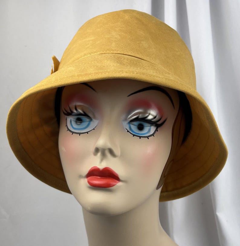 Puede incluir: Un sombrero de pescador color amarillo mostaza con un ala ancha inclinada hacia abajo y un peque&ntilde;o detalle de lazo decorativo en el lateral. El sombrero est&aacute; confeccionado en una tela suave similar a la gamuza y se exhibe en un maniqu&iacute;.