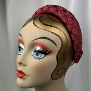 May include: A red headband with a repeating floral pattern. The headband is on a mannequin head with painted features, including blue eyes, red lips, and gold-painted hair.
