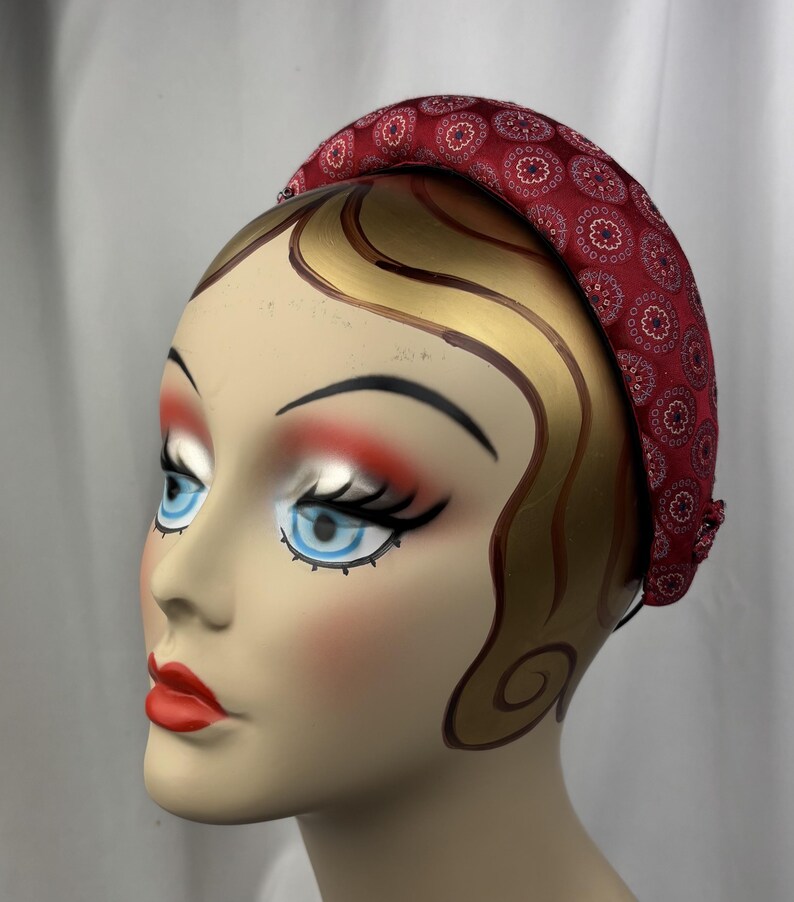 May include: A red headband with a repeating floral pattern. The headband is on a mannequin head with painted features, including blue eyes, red lips, and gold-painted hair.