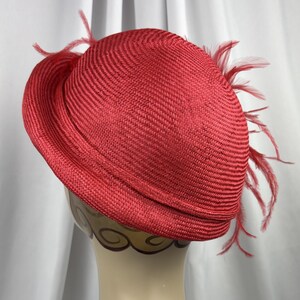 May include: A vibrant red straw hat featuring a rounded crown and a structured brim. The hat is adorned with delicate, matching red feathers that extend from the side, adding texture and movement to the woven material of the headwear.