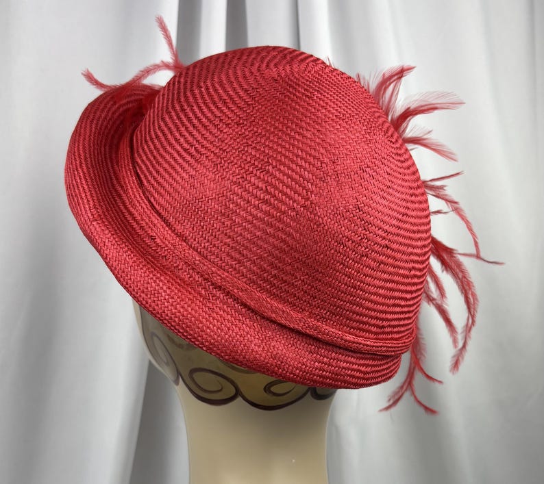 May include: A vibrant red straw hat featuring a rounded crown and a structured brim. The hat is adorned with delicate, matching red feathers that extend from the side, adding texture and movement to the woven material of the headwear.