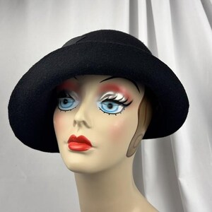 May include: A solid black felt cloche hat with a rounded crown and a slightly downturned brim. The hat features a simple, clean design with a smooth texture, providing a classic look that sits securely on the head of the mannequin display.