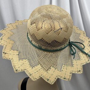 May include: A woven straw sun hat with a wide, scalloped brim. The hat features a green ribbon around the crown and a matching bow. The brim is trimmed with a decorative pattern. The hat is approximately 15 inches in diameter.