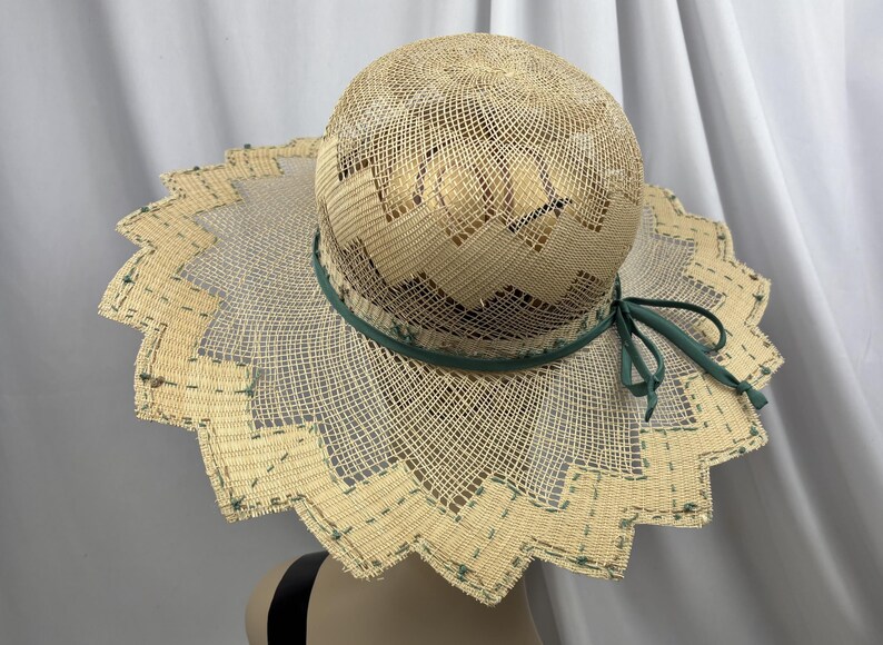 May include: A woven straw sun hat with a wide, scalloped brim. The hat features a green ribbon around the crown and a matching bow. The brim is trimmed with a decorative pattern. The hat is approximately 15 inches in diameter.