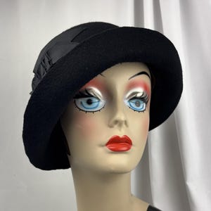 May include: A black felt cloche hat featuring a wide, downward-sloping brim and a decorative black grosgrain ribbon bow detail on the side. The hat is constructed from a solid black material with a smooth texture and a structured, rounded crown.