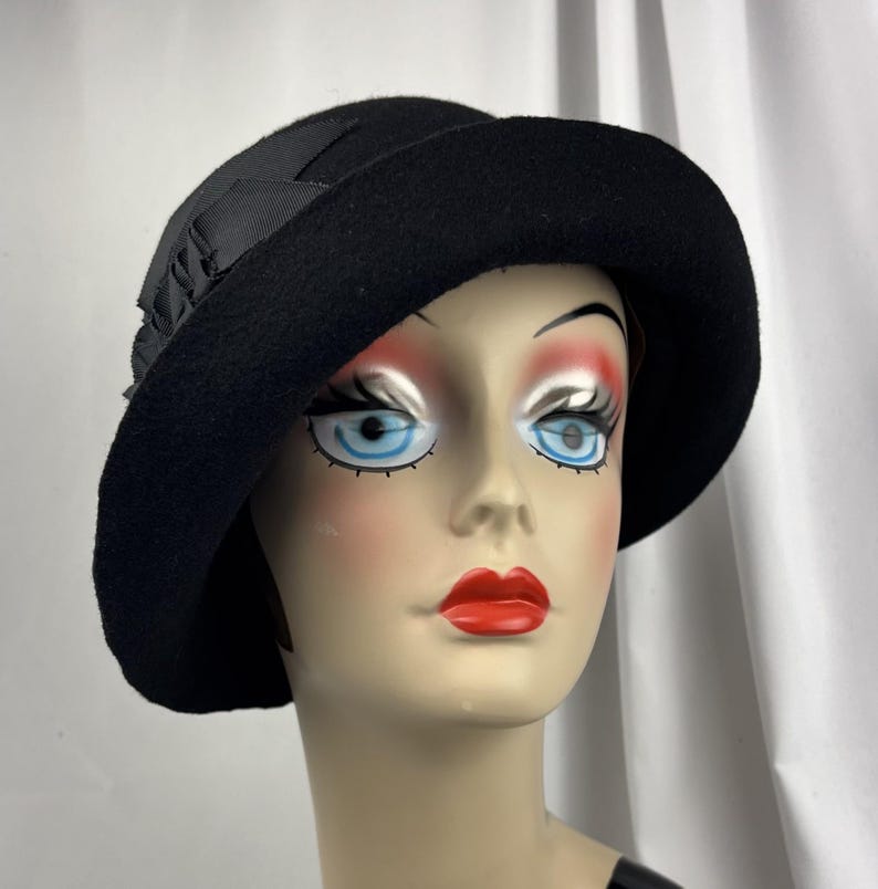May include: A black felt cloche hat featuring a wide, downward-sloping brim and a decorative black grosgrain ribbon bow detail on the side. The hat is constructed from a solid black material with a smooth texture and a structured, rounded crown.