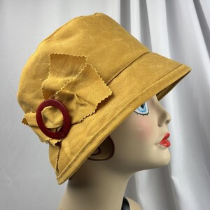Puede incluir: Un sombrero de pescador color amarillo mostaza con un detalle lateral compuesto por un lazo de tela con bordes dentados y un anillo circular de color rojo oscuro. El sombrero tiene una copa estructurada y un ala inclinada hacia abajo.