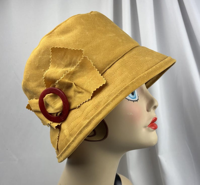 Puede incluir: Un sombrero de pescador color amarillo mostaza con un detalle lateral compuesto por un lazo de tela con bordes dentados y un anillo circular de color rojo oscuro. El sombrero tiene una copa estructurada y un ala inclinada hacia abajo.