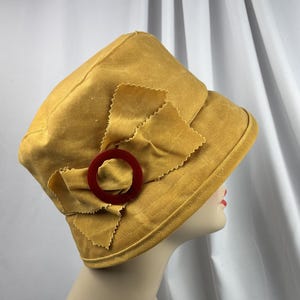 Puede incluir: Un sombrero de color amarillo mostaza con un detalle de lazo de tela en el lateral. El lazo est&aacute; adornado con un anillo circular de pl&aacute;stico rojo oscuro. El sombrero tiene una copa estructurada y un ala inclinada, hecho de tela lisa.