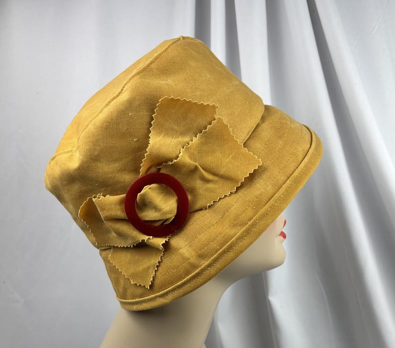 Puede incluir: Un sombrero de color amarillo mostaza con un detalle de lazo de tela en el lateral. El lazo est&aacute; adornado con un anillo circular de pl&aacute;stico rojo oscuro. El sombrero tiene una copa estructurada y un ala inclinada, hecho de tela lisa.