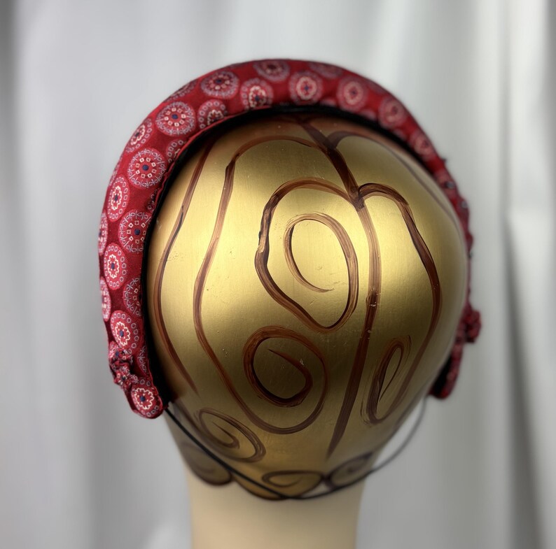 May include: A red headband with a repeating circular pattern in white and pink. The headband is on a gold-colored head form with brown swirl designs. The headband is a fashion accessory.