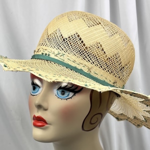 May include: A straw hat with a wide brim and a decorative green ribbon. The hat features a woven design and a scalloped edge. The hat is on a mannequin head with painted features, including blue eyes and red lips.