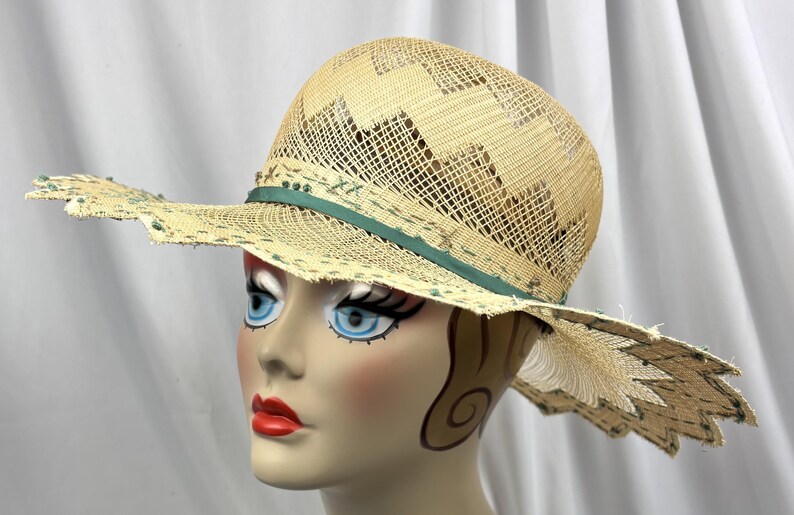 May include: A straw hat with a wide brim and a decorative green ribbon. The hat features a woven design and a scalloped edge. The hat is on a mannequin head with painted features, including blue eyes and red lips.