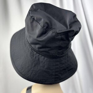Pu&ograve; includere: Un cappello a secchiello nero con tesa larga. Il cappello &egrave; realizzato in un tessuto liscio, possibilmente impermeabile. La corona del cappello &egrave; leggermente arricciata e la tesa &egrave; delicatamente curva. Il cappello &egrave; di colore nero pieno.