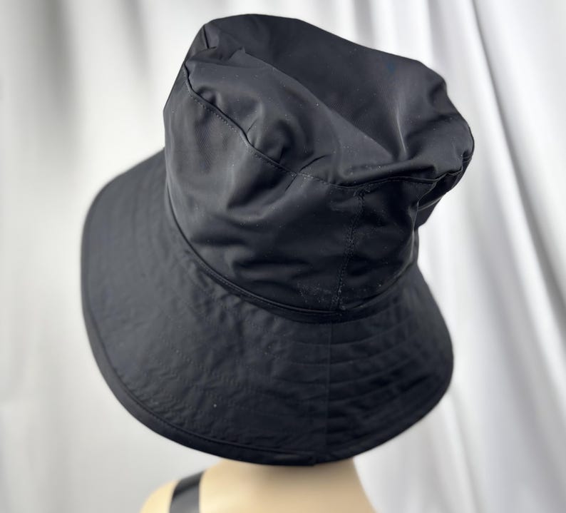Pu&ograve; includere: Un cappello a secchiello nero con tesa larga. Il cappello &egrave; realizzato in un tessuto liscio, possibilmente impermeabile. La corona del cappello &egrave; leggermente arricciata e la tesa &egrave; delicatamente curva. Il cappello &egrave; di colore nero pieno.