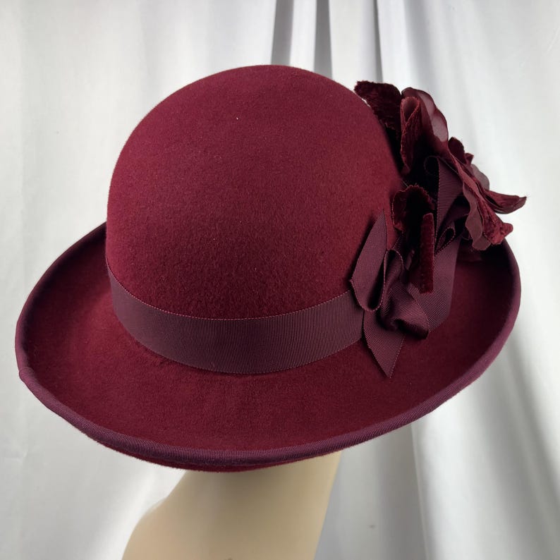 May include: A deep burgundy felt cloche hat featuring a matching grosgrain ribbon band around the crown. The side of the hat is adorned with a cluster of layered fabric flowers in a coordinating dark red shade, finished with a bound brim edge.
