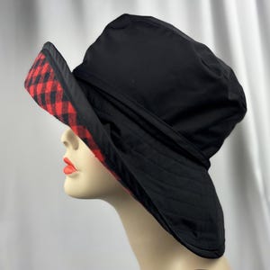Pu&ograve; includere: Cappello da pescatore nero con tesa a scacchi rossi e neri. Il cappello &egrave; realizzato in materiale trapuntato e ha un'ampia tesa. Il cappello &egrave; mostrato su una testa di manichino.