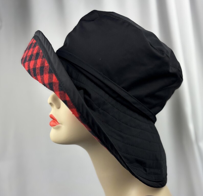 Pu&ograve; includere: Cappello da pescatore nero con tesa a scacchi rossi e neri. Il cappello &egrave; realizzato in materiale trapuntato e ha un'ampia tesa. Il cappello &egrave; mostrato su una testa di manichino.