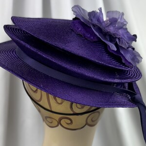May include: A layered, purple hat with a wide brim and a decorative flower on top. The hat has a dark purple ribbon and is displayed on a mannequin head. The hat is made of a woven material.