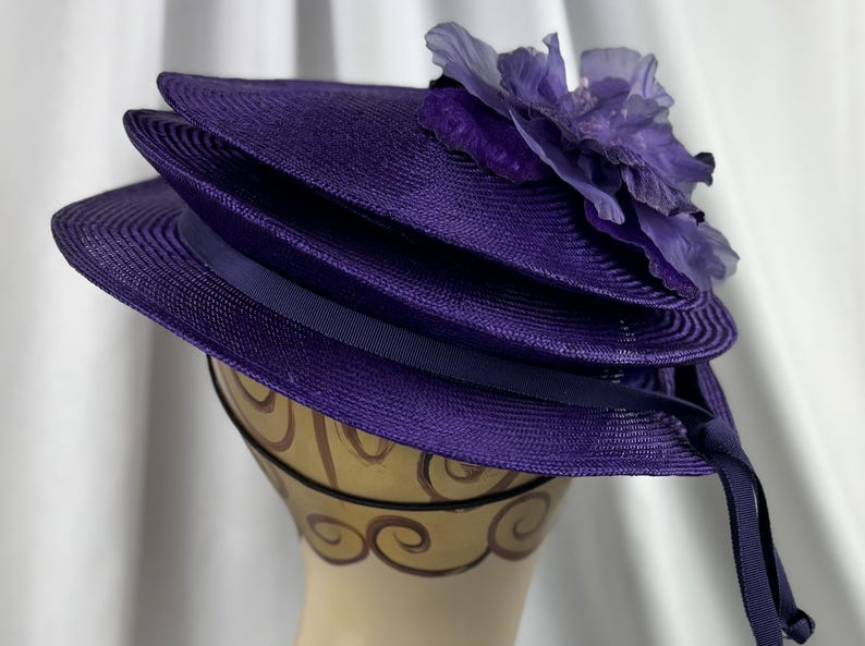May include: A layered, purple hat with a wide brim and a decorative flower on top. The hat has a dark purple ribbon and is displayed on a mannequin head. The hat is made of a woven material.