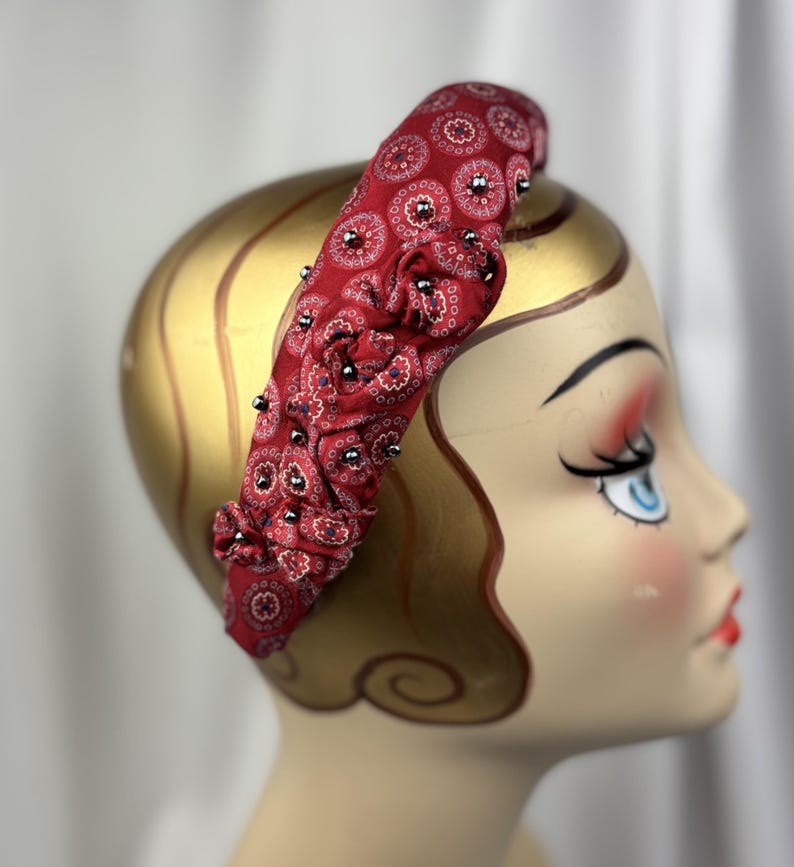 May include: A red headband with a paisley pattern and small silver beads. The fabric is gathered in sections, creating a textured look. The headband is displayed on a mannequin head.