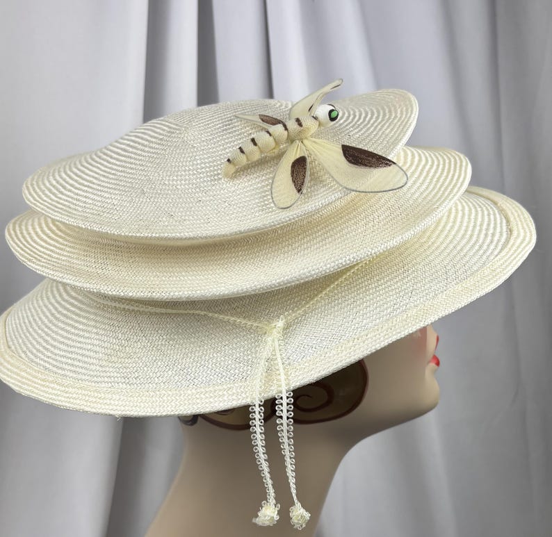 May include: A cream-colored multi-tiered straw hat featuring a prominent dragonfly ornament on the top layer. The dragonfly has translucent wings with dark brown accents and a green eye. A thin, decorative cord with small beaded tassels hangs down.