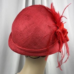 May include: A bright red cloche hat constructed from a woven straw-like material. The hat features a rounded crown and a narrow, slightly upturned brim. A decorative red fabric flower accented with long, thin red feathers adorns the side.