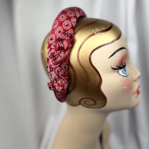 May include: A red fabric headband with a paisley pattern and small black embellishments. The headband is wrapped around a mannequin head with gold-painted skin and a vintage-style face.