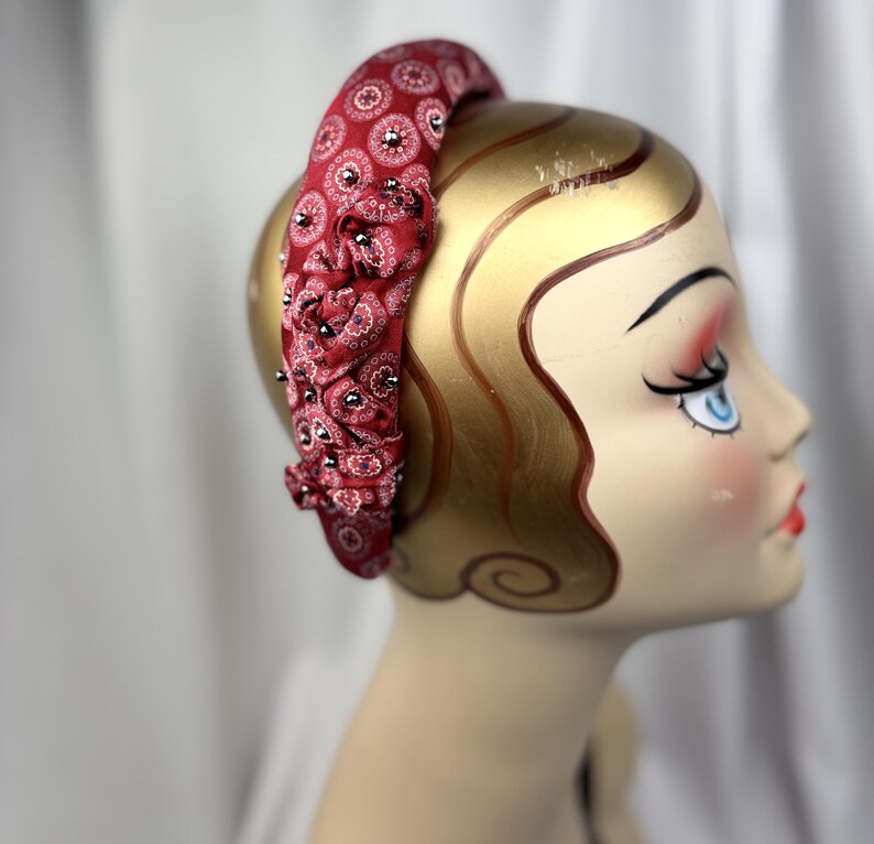 May include: A red fabric headband with a paisley pattern and small black embellishments. The headband is wrapped around a mannequin head with gold-painted skin and a vintage-style face.