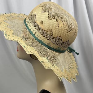May include: A wide-brimmed straw hat with a decorative green ribbon and trim. The hat features a unique open weave design with geometric cutouts and a scalloped edge. The hat is displayed on a mannequin head.