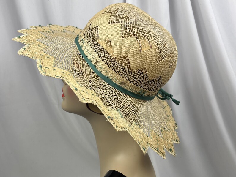 May include: A wide-brimmed straw hat with a decorative green ribbon and trim. The hat features a unique open weave design with geometric cutouts and a scalloped edge. The hat is displayed on a mannequin head.
