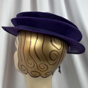 May include: A deep purple hat with a wide brim and a structured, layered design. The hat features a dark purple band and a textured weave, suggesting a formal or vintage style. The hat is displayed on a mannequin head.