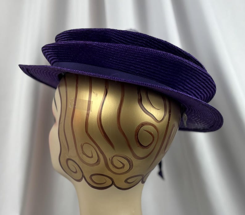 May include: A deep purple hat with a wide brim and a structured, layered design. The hat features a dark purple band and a textured weave, suggesting a formal or vintage style. The hat is displayed on a mannequin head.