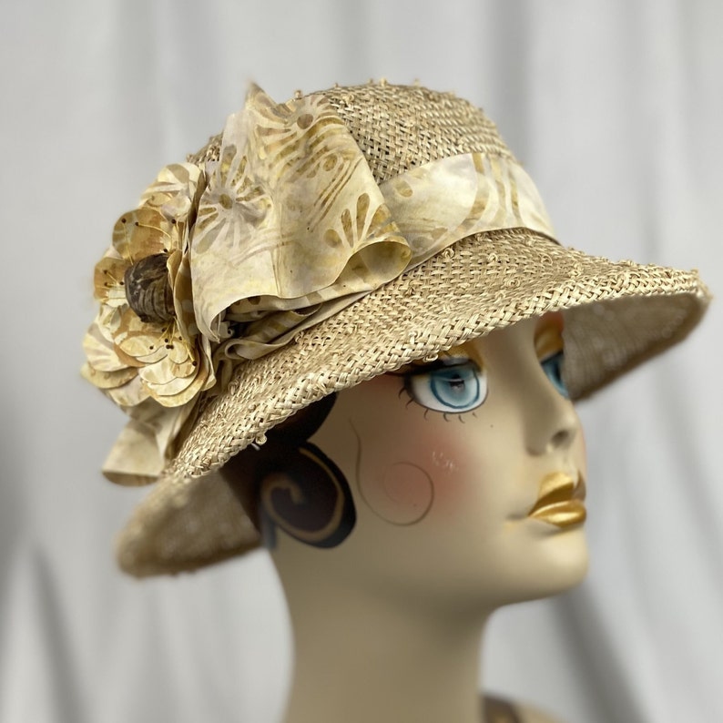 Puede incluir: Un sombrero de paja de ala ancha con una gran flor de tela beige como adorno. El sombrero est&aacute; decorado con una cinta beige.