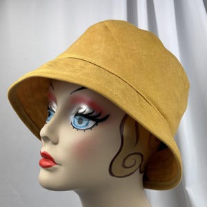 Puede incluir: Un sombrero de pescador color amarillo mostaza con un ala ancha inclinada hacia abajo y una copa redondeada. El sombrero se muestra sobre una cabeza de maniqu&iacute; con rasgos faciales estilizados, incluyendo ojos azules, pesta&ntilde;as oscuras y labios rojos brillantes.