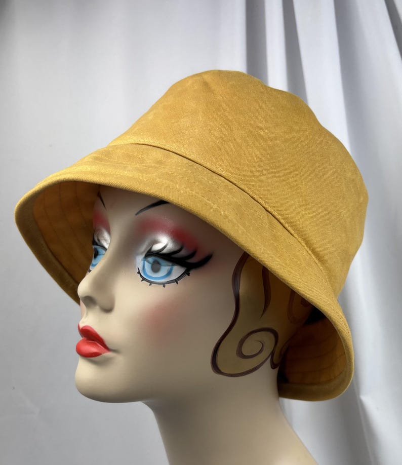 Puede incluir: Un sombrero de pescador color amarillo mostaza con un ala ancha inclinada hacia abajo y una copa redondeada. El sombrero se muestra sobre una cabeza de maniqu&iacute; con rasgos faciales estilizados, incluyendo ojos azules, pesta&ntilde;as oscuras y labios rojos brillantes.