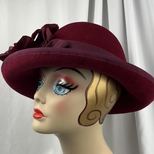 May include: A deep burgundy felt hat featuring a wide brim and a matching grosgrain ribbon band. The hat is adorned with a cluster of fabric flowers in the same dark red tone, positioned on the side of the crown above the brim.