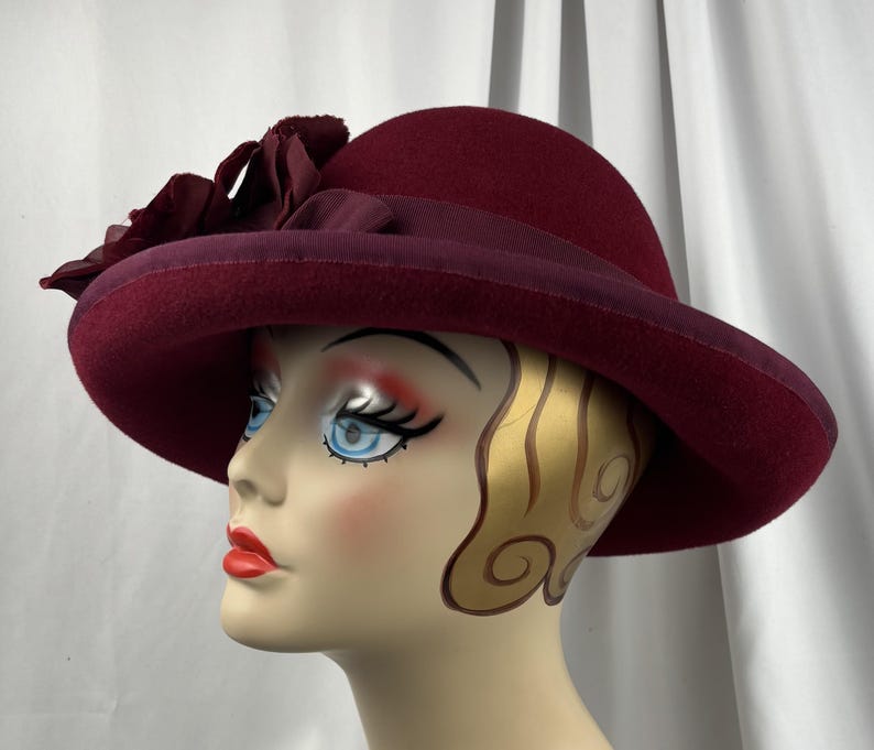 May include: A deep burgundy felt hat featuring a wide brim and a matching grosgrain ribbon band. The hat is adorned with a cluster of fabric flowers in the same dark red tone, positioned on the side of the crown above the brim.