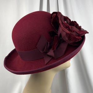May include: A deep burgundy felt cloche hat featuring a wide grosgrain ribbon band and a large, layered fabric flower accent on the side. The hat has a rounded crown and a short, slightly upturned brim, finished with matching trim along the edge.