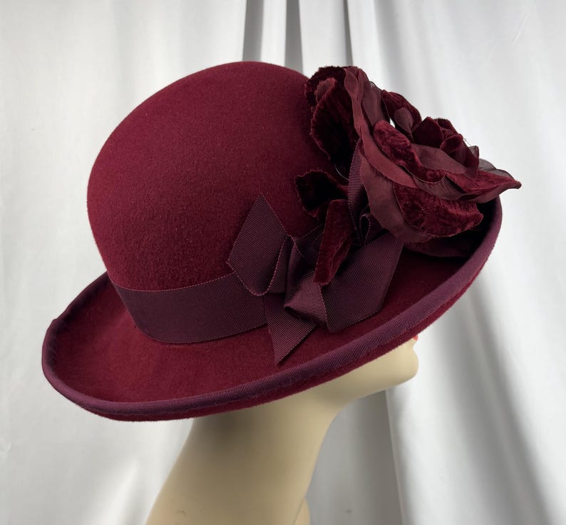 May include: A deep burgundy felt cloche hat featuring a wide grosgrain ribbon band and a large, layered fabric flower accent on the side. The hat has a rounded crown and a short, slightly upturned brim, finished with matching trim along the edge.