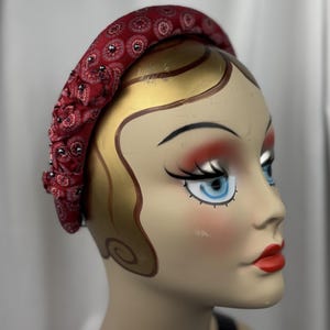 May include: A red fabric headband with a paisley pattern and small, dark embellishments. The headband is displayed on a mannequin head with painted features, including red lips and blue eyes. The headband is a fashion accessory.