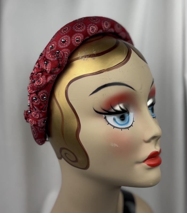 May include: A red fabric headband with a paisley pattern and small, dark embellishments. The headband is displayed on a mannequin head with painted features, including red lips and blue eyes. The headband is a fashion accessory.