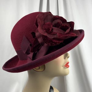 May include: A deep burgundy felt hat featuring a wide brim and a matching grosgrain ribbon band. The hat is adorned with a large, layered velvet flower decoration in the same dark red tone, positioned on the side above the brim.