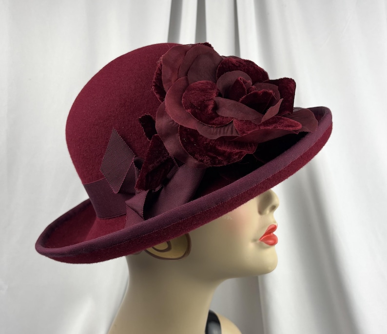 May include: A deep burgundy felt hat featuring a wide brim and a matching grosgrain ribbon band. The hat is adorned with a large, layered velvet flower decoration in the same dark red tone, positioned on the side above the brim.