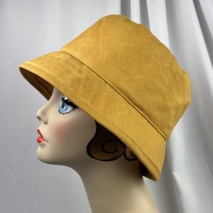Puede incluir: Un sombrero de pescador color amarillo mostaza con copa redondeada y ala inclinada hacia abajo. El sombrero presenta costuras visibles a lo largo del ala y est&aacute; confeccionado con una tela de color s&oacute;lido con textura mate, sobre una cabeza de maniqu&iacute;.
